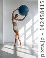 Slim female fitness enthusiast in white sports bra and beige shorts holds a blue exercise ball while posing in a bright, sunlit studio with geometric shadows 142458415