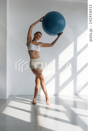 Slim female fitness enthusiast in white sports bra and beige shorts holds a blue exercise ball while posing in a bright, sunlit studio with geometric shadows Slim female fitness enthusiast in white sports bra and beige shorts holds a blue exercise ball while posing in a bright, sunlit studio with geometric shadows 142458415