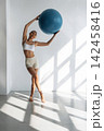 Slim female fitness model in white sports bra and beige shorts poses with a blue exercise ball in a bright, modern studio with sunlight streaming through large windows 142458416