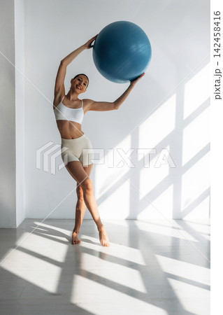 Slim female fitness model in white sports bra and beige shorts poses with a blue exercise ball in a bright, modern studio with sunlight streaming through large windows Slim female fitness model in white sports bra and beige shorts poses with a blue exercise ball in a bright, modern studio with sunlight streaming through large windows 142458416