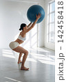 Female fitness enthusiast performing a squat while holding a large blue exercise ball in a bright, modern gym studio with large windows and wooden flooring 142458418