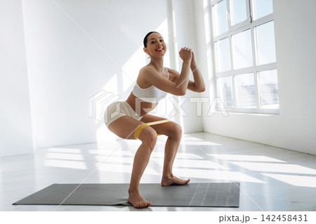 Slim female fitness model performing squats with resistance band in a bright, modern gym studio with large windows and natural light illuminating the space Slim female fitness model performing squats with resistance band in a bright, modern gym studio with large windows and natural light illuminating the space 142458431