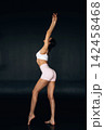 Slim female dancer in white crop top and pink shorts poses gracefully with arms raised against a dark background, showcasing her ideal curves and athletic physique 142458468