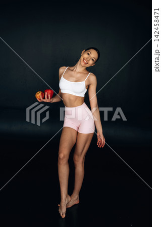 Female model with long hair wearing white crop top and pink shorts holds red apples while posing against a dark background in a studio setting 142458471