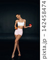 Slim female model in white crop top and pink shorts holding red apple and orange, posing against a dark background with a confident expression 142458474