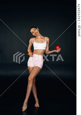Slim female model in white crop top and pink shorts holding red apple and orange, posing against a dark background with a confident expression Slim female model in white crop top and pink shorts holding red apple and orange, posing against a dark background with a confident expression 142458474