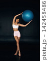 Slim female fitness model in white sports bra and pink shorts poses with a blue exercise ball against a dark background, showcasing a healthy physique and athleticism 142458486