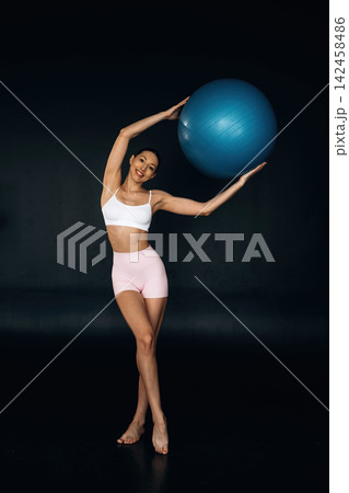 Slim female fitness model in white sports bra and pink shorts poses with a blue exercise ball against a dark background, showcasing a healthy physique and athleticism Slim female fitness model in white sports bra and pink shorts poses with a blue exercise ball against a dark background, showcasing a healthy physique and athleticism 142458486