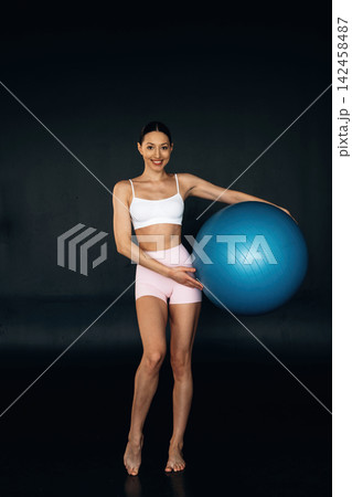 Slim female fitness model in white sports bra and pink shorts poses with a blue exercise ball against a dark background in a studio setting 142458487