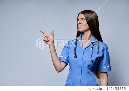 Smiling female nurse pointing finger to side copy space isolated against grey background Smiling female nurse pointing finger to side copy space isolated against grey background 142458876
