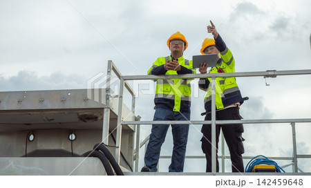 Two Asian male electrical engineers wearing hardhats and safety vests use tablet and walkie-talkie while pointing ahead on a high platform structure. 142459648
