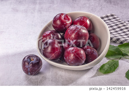 Fresh plums in ceramic bowl with green leaves and checkered cloth on gray surface Fresh plums in ceramic bowl with green leaves and checkered cloth on gray surface 142459916
