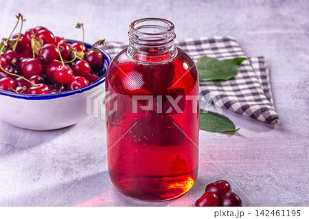 Glass bottle filled with red cherry liquid beside bowl of fresh cherries on textured surface Glass bottle filled with red cherry liquid beside bowl of fresh cherries on textured surface 142461195