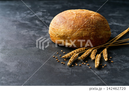 Round loaf of wheat rustic bread with sourdough and ears of wheat on dark kitchen table, selective focus. Round loaf of wheat rustic bread with sourdough and ears of wheat on dark kitchen table, selective focus. 142462720