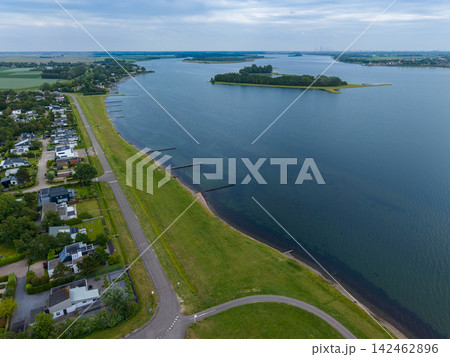 High angle wide horizontal aerial drone photograph of coastal road, grass dike, waterfront houses, Lake Veere, and green island in Ruiterplaat, Zeeland, Netherlands on overcast summer day 142462896