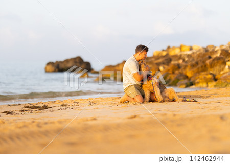 Young man kneeling on sandy beach embraces golden retriever dog with rocky shoreline and calm ocean waves visible in the background during sunset. Airedale Terrier Young man kneeling on sandy beach embraces golden retriever dog with rocky shoreline and calm ocean waves visible in the background during sunset. Airedale Terrier 142462944
