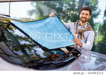 A smiling caucasian man holds a blue microfiber cloth to clean the windshield of a car outdoors A smiling caucasian man holds a blue microfiber cloth to clean the windshield of a car outdoors 142463123