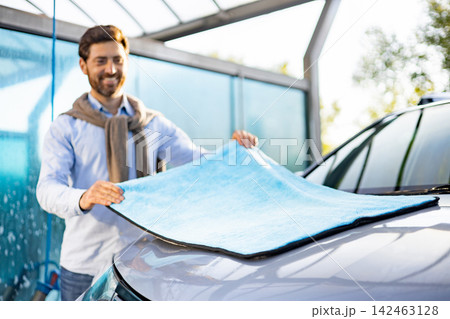 A smiling man holds a blue microfiber towel over the hood of a car at a car wash A smiling man holds a blue microfiber towel over the hood of a car at a car wash 142463128