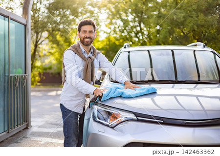 A smiling Caucasian man is meticulously polishing the hood of his silver car with a bright blue microfiber cloth on a sunny day 142463362