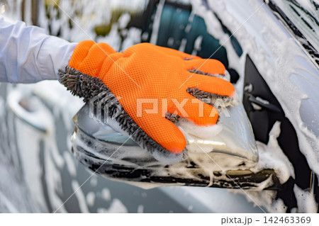 A person wearing an orange microfiber wash mitt cleans a soapy car side mirror with suds and foam A person wearing an orange microfiber wash mitt cleans a soapy car side mirror with suds and foam 142463369