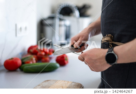 Man In A Kitchen Apron Honing A Knife Blade In Preparation For A Meal Man In A Kitchen Apron Honing A Knife Blade In Preparation For A Meal 142464267