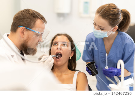 Girl is sitting in armchair during dental treatment at dentist appointment. Male doctor performs procedure of restoration and improvement of teeth, uses technological device 142467250