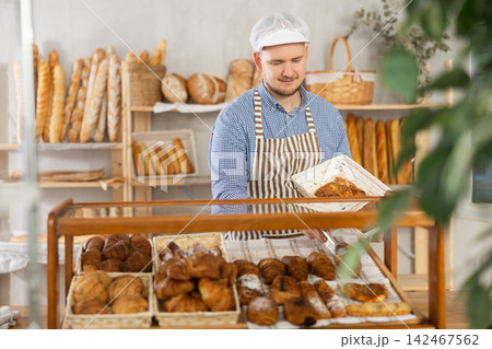Man employee puts croissants in window, arranges display of goods at bakery. Man employee puts croissants in window, arranges display of goods at bakery. 142467562