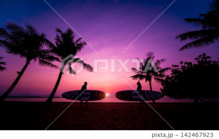 Silhouette of surfers couple holding long surf boards at sunset on tropical beach Silhouette of surfers couple holding long surf boards at sunset on tropical beach 142467923