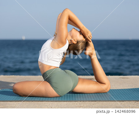 Flexible fit woman doing yoga on the seashore, performing seated back-bending asana Rajakapotasana, or King Pigeon Pose 142468096