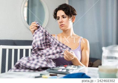 Young woman repairing plaid shirt with needle and thread 142468302