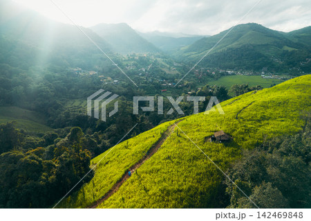 Aerial view of a scenic dirt trail leading up a vibrant green grassy hill in a lush mountain valley. 142469848