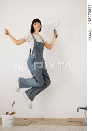 A joyful woman in denim overalls leaps mid-air with a paint roller, ready for home renovation 142470898