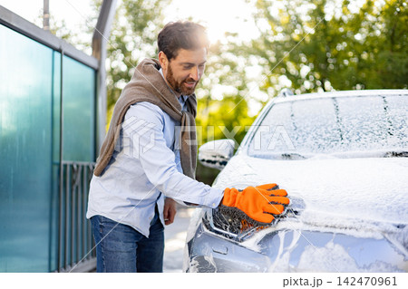 A smiling man wearing an orange glove washes his car with soap suds on a sunny day 142470961