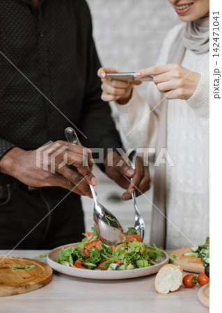 A man and woman are preparing a fresh salad, with the woman capturing the moment on her smartphone 142471041