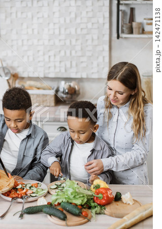 A mother and her two sons are happily preparing a healthy meal in a bright, modern kitchen, chopping fresh vegetables for a salad A mother and her two sons are happily preparing a healthy meal in a bright, modern kitchen, chopping fresh vegetables for a salad 142471138
