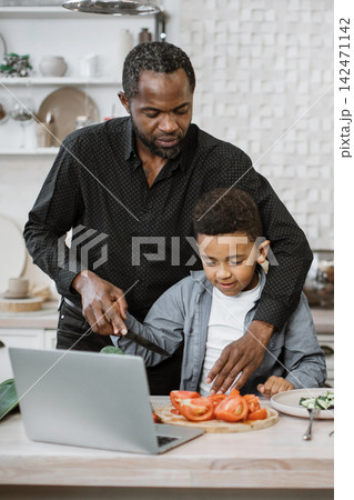 A father guides his young son as they prepare vegetables in a modern kitchen, with a laptop open for a recipe or video call 142471142