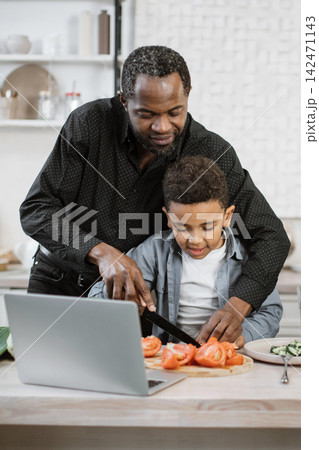 A father guides his young son as they chop tomatoes in the kitchen, following a recipe on a laptop 142471143