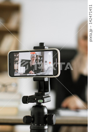 A woman wearing a hijab is recording herself on a smartphone mounted on a tripod, likely for a vlog or online class A woman wearing a hijab is recording herself on a smartphone mounted on a tripod, likely for a vlog or online class 142471451