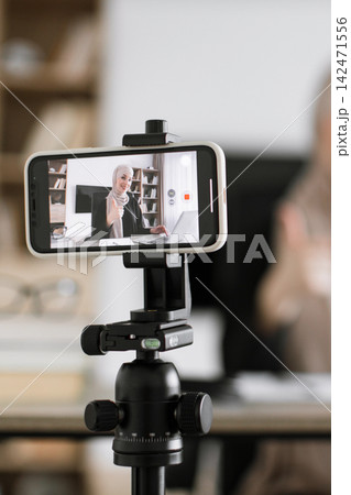 A woman wearing a hijab records herself giving a thumbs-up gesture while working on her laptop, with the recording displayed on a smartphone mounted on a tripod A woman wearing a hijab records herself giving a thumbs-up gesture while working on her laptop, with the recording displayed on a smartphone mounted on a tripod 142471556