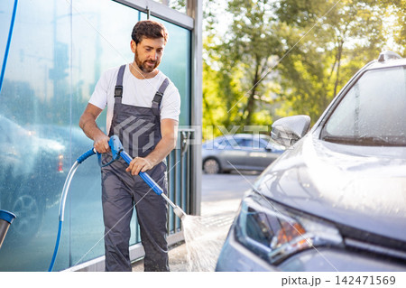 A man in overalls uses a high-pressure washer to clean a car at an outdoor car wash station A man in overalls uses a high-pressure washer to clean a car at an outdoor car wash station 142471569