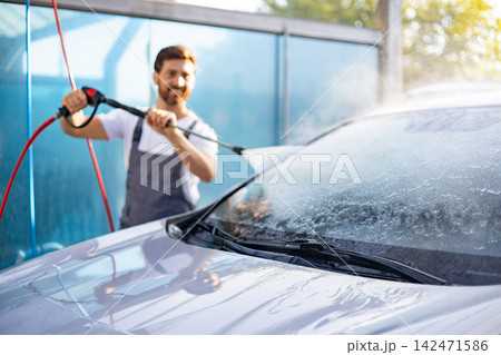 A smiling Caucasian man in overalls uses a high-pressure washer to clean the windshield of a car at an outdoor car wash station A smiling Caucasian man in overalls uses a high-pressure washer to clean the windshield of a car at an outdoor car wash station 142471586
