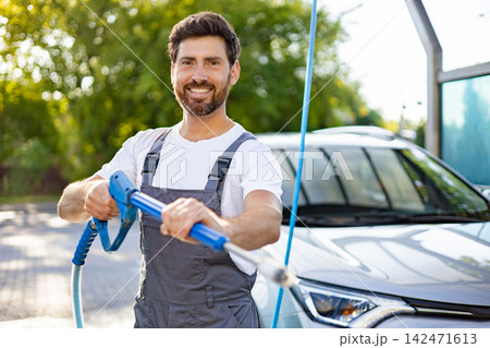 A smiling Caucasian man in overalls holds a pressure washer wand, ready to clean a car at an outdoor wash station A smiling Caucasian man in overalls holds a pressure washer wand, ready to clean a car at an outdoor wash station 142471613