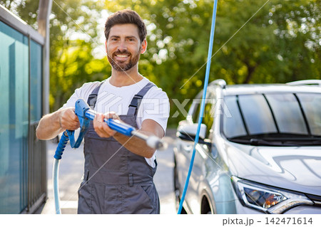 A smiling caucasian man in overalls holds a pressure washer wand, ready to clean a silver car outdoors on a sunny day A smiling caucasian man in overalls holds a pressure washer wand, ready to clean a silver car outdoors on a sunny day 142471614