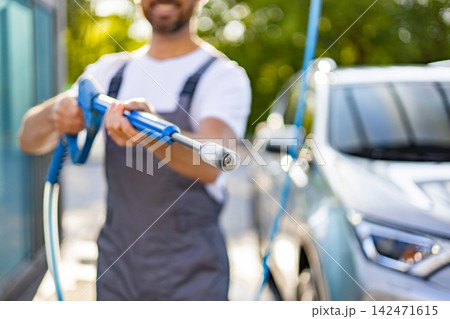 A smiling man in overalls uses a high-pressure washer to clean a car outdoors on a sunny day 142471615