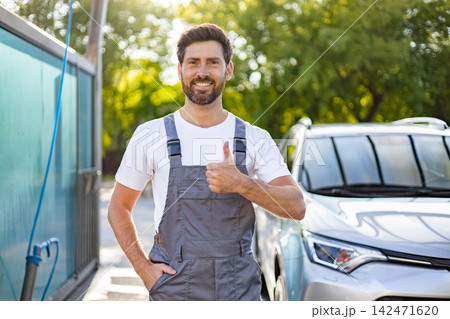 A smiling caucasian man in overalls gives a thumbs up gesture while standing next to a car at a car wash 142471620