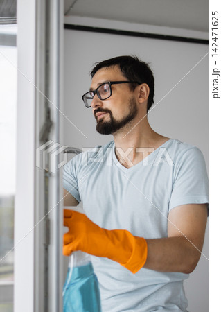 A man wearing orange gloves and glasses sprays a window with a cleaning solution, preparing to wash it 142471625