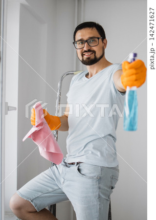 A smiling man wearing glasses and orange gloves holds a pink cloth and a spray bottle while standing on a ladder A smiling man wearing glasses and orange gloves holds a pink cloth and a spray bottle while standing on a ladder 142471627