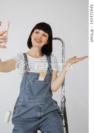 A young woman in overalls smiles while taking a selfie with her phone, a paintbrush tucked into her pocket and a roller visible A young woman in overalls smiles while taking a selfie with her phone, a paintbrush tucked into her pocket and a roller visible 142471648