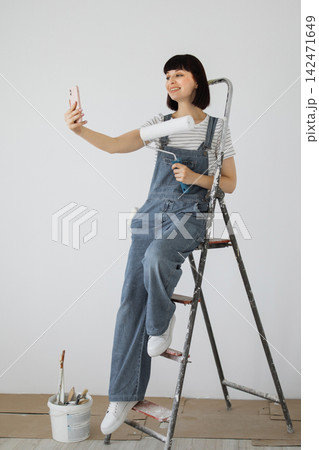 A young woman in denim overalls sits on a ladder holding a paint roller and a smartphone, taking a selfie during home renovation A young woman in denim overalls sits on a ladder holding a paint roller and a smartphone, taking a selfie during home renovation 142471649