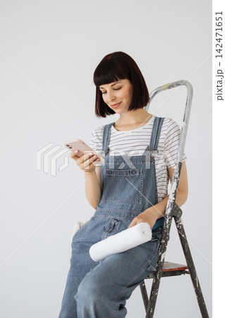 A woman in denim overalls sits on a ladder, holding a phone and a paint roller, looking at her device with a smile A woman in denim overalls sits on a ladder, holding a phone and a paint roller, looking at her device with a smile 142471651